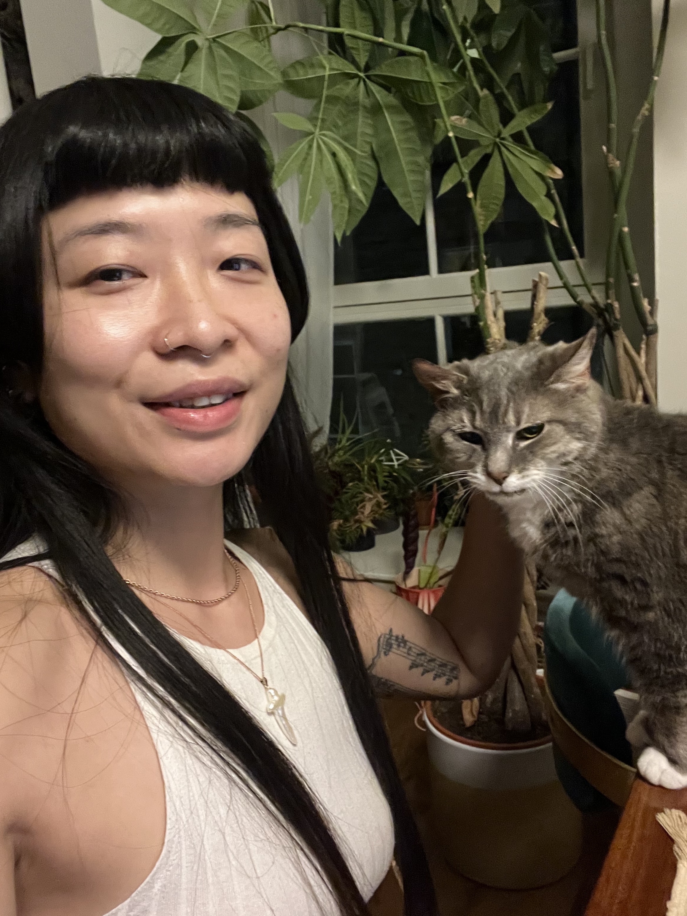 A Chinese woman with a blunt fringe in her mid-thirties pets a grey tabby cat whilst smiling into the camera. 