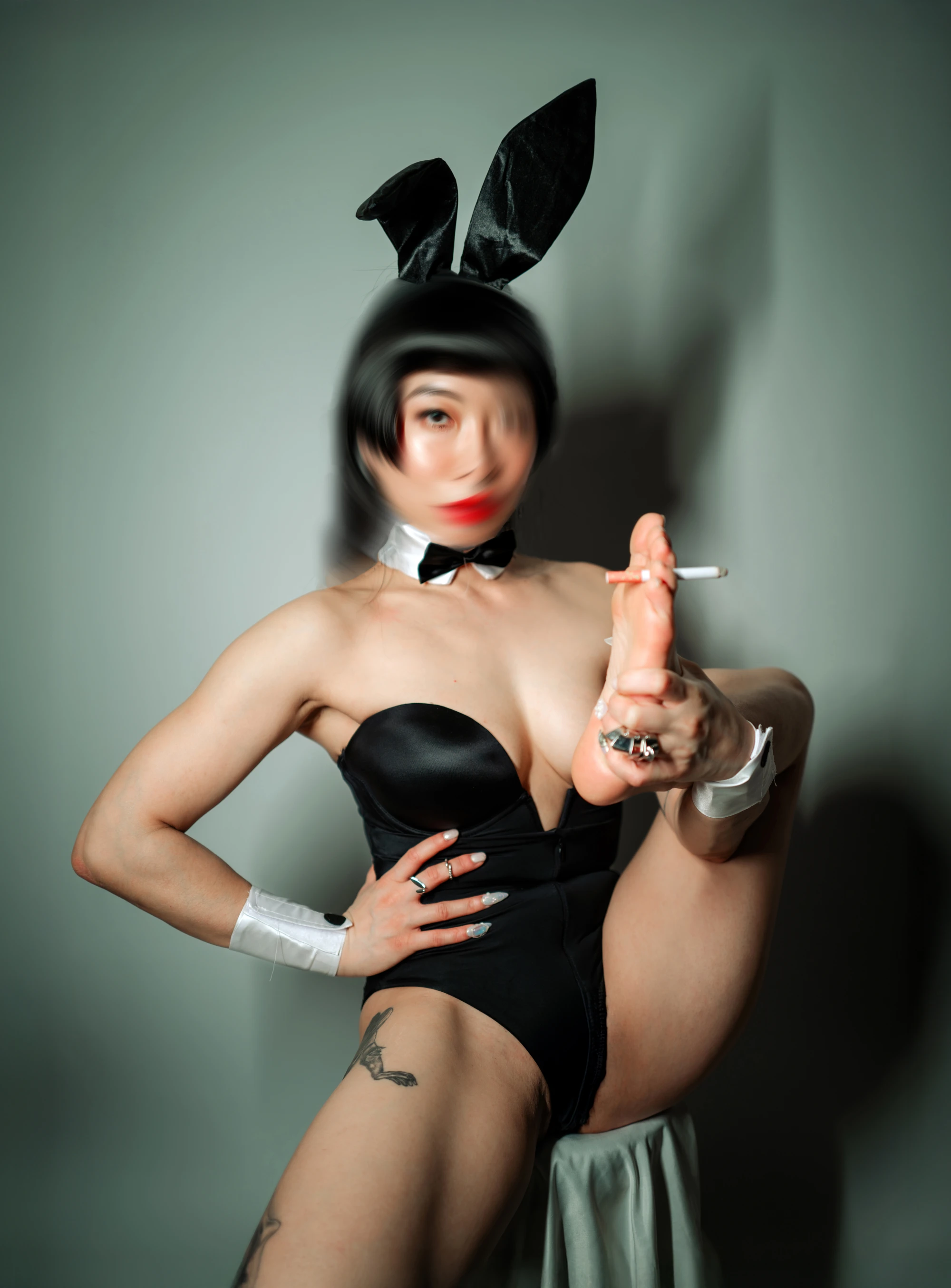 a woman with a semi-blurred face, wearing a playboy bunny costume, sits on top of a fabric draped stool holding up a cigarette with her foot.