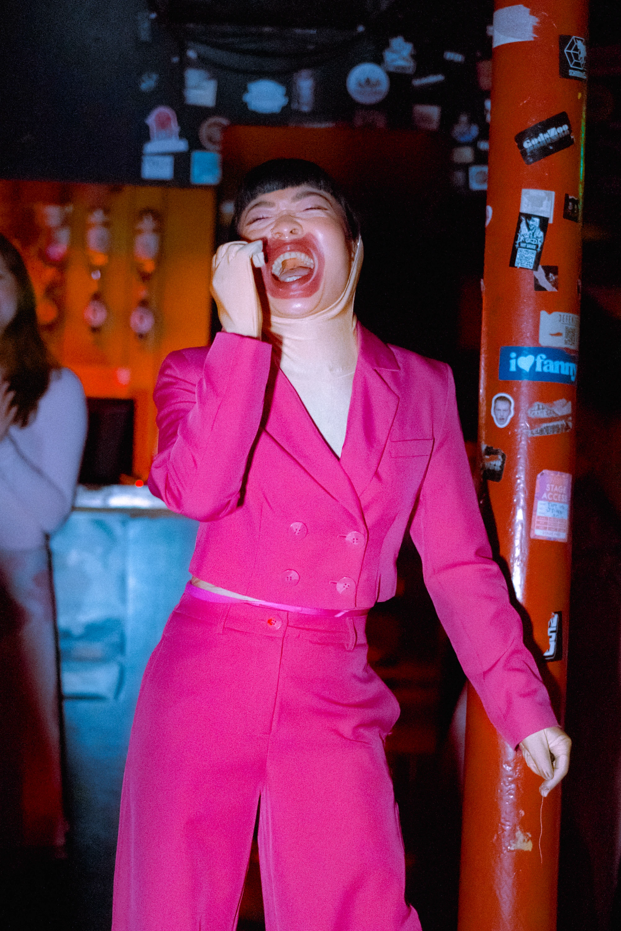 a woman wearing a pink suit over a flesh covered bodysuit applies lipstick all over her face.