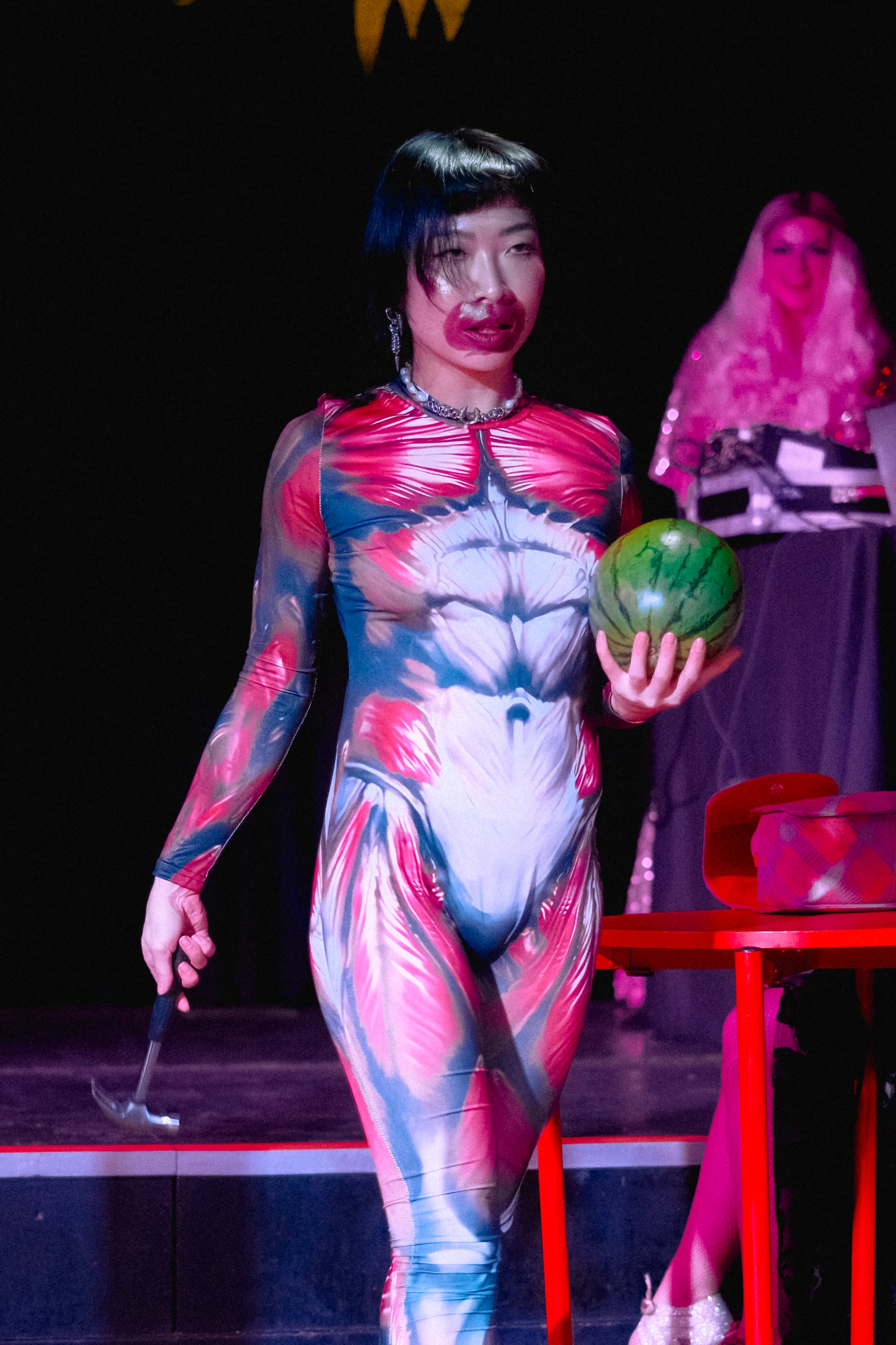 a woman wearing an anatomical muscle bodysuit is holding a watermelon and a hammer.