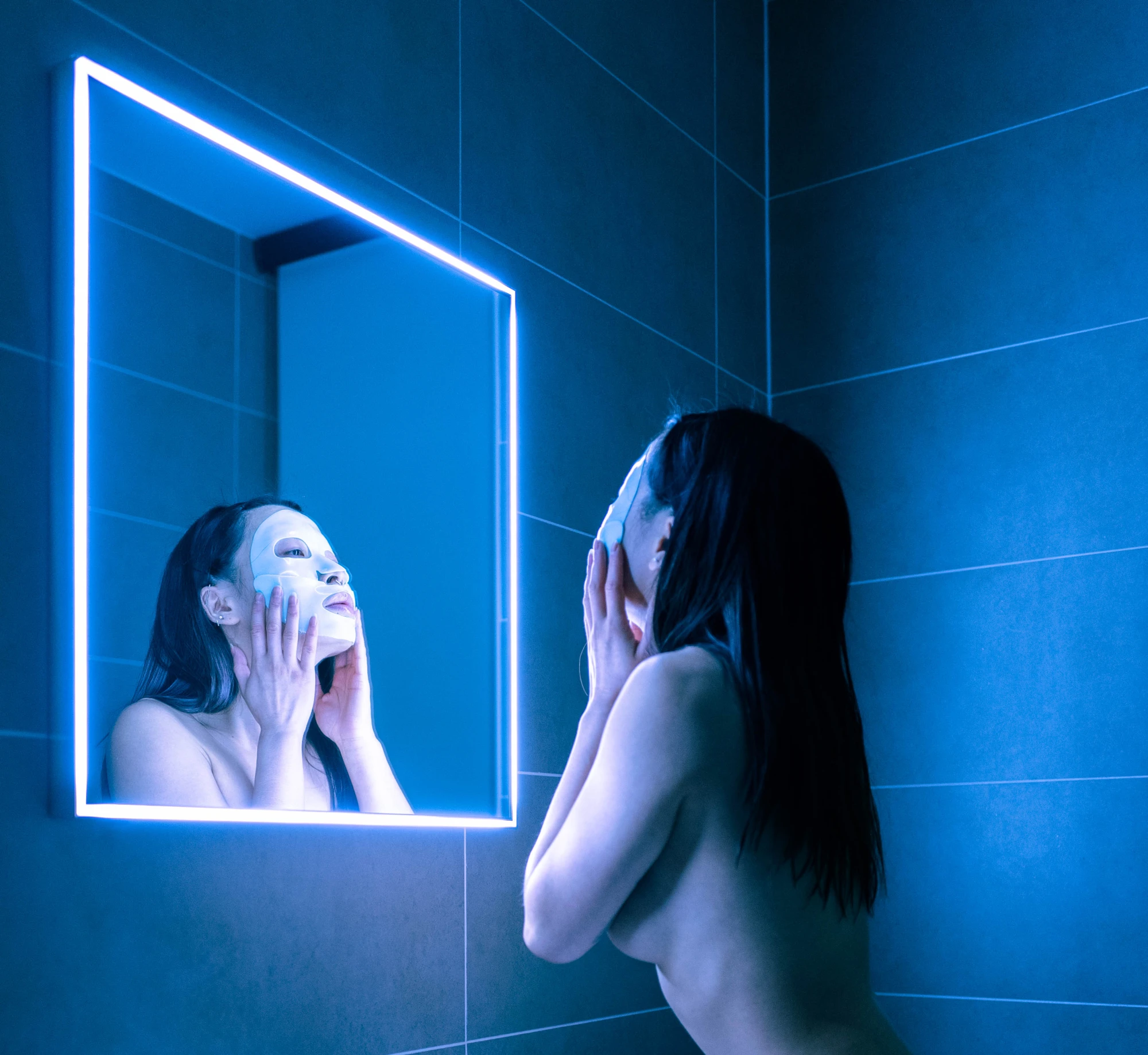 a woman wearing a rubber face mask stands in front of a neon lit mirror.