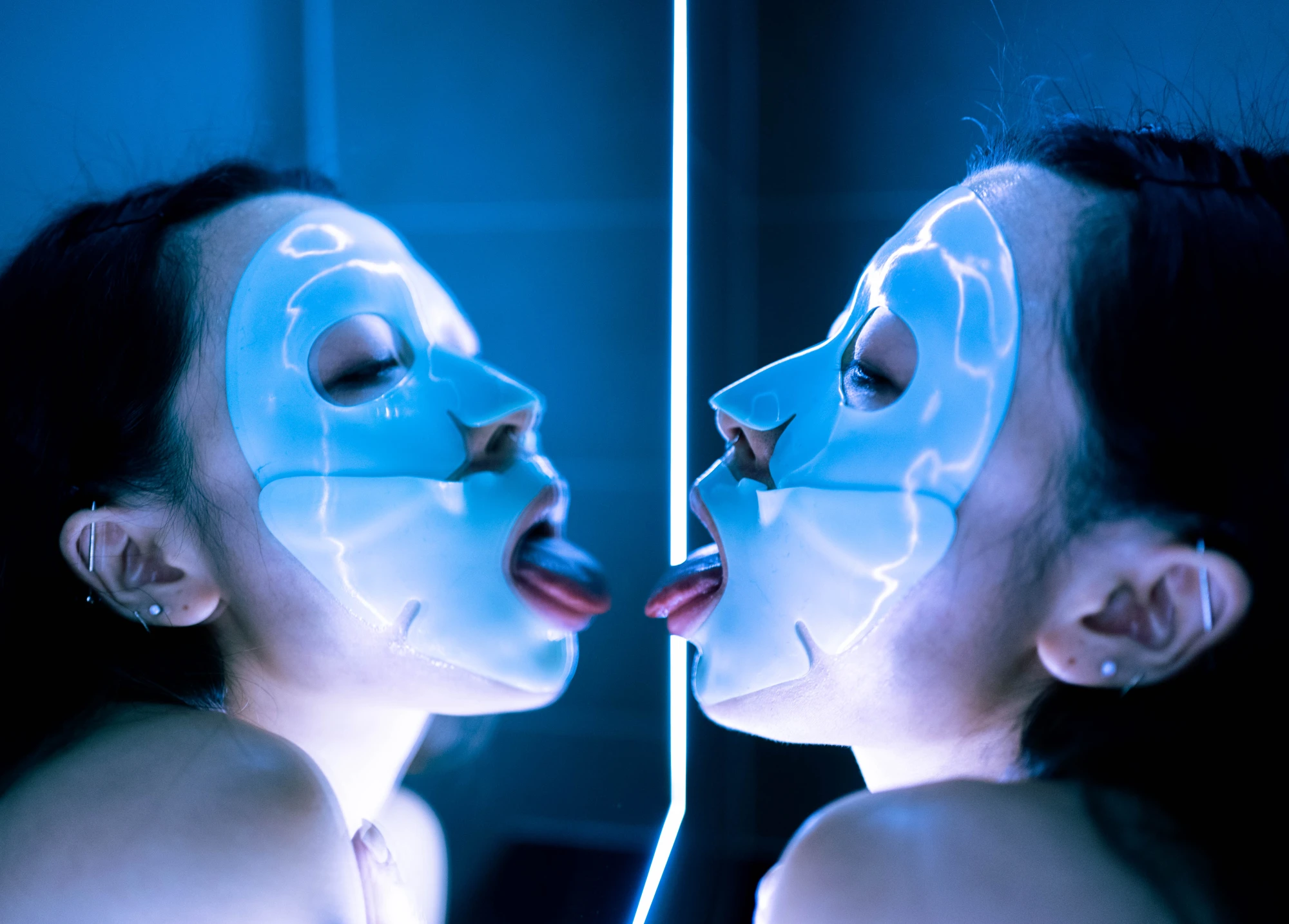 a woman wearing a rubber face mask licks her reflection in a neon lit mirror.