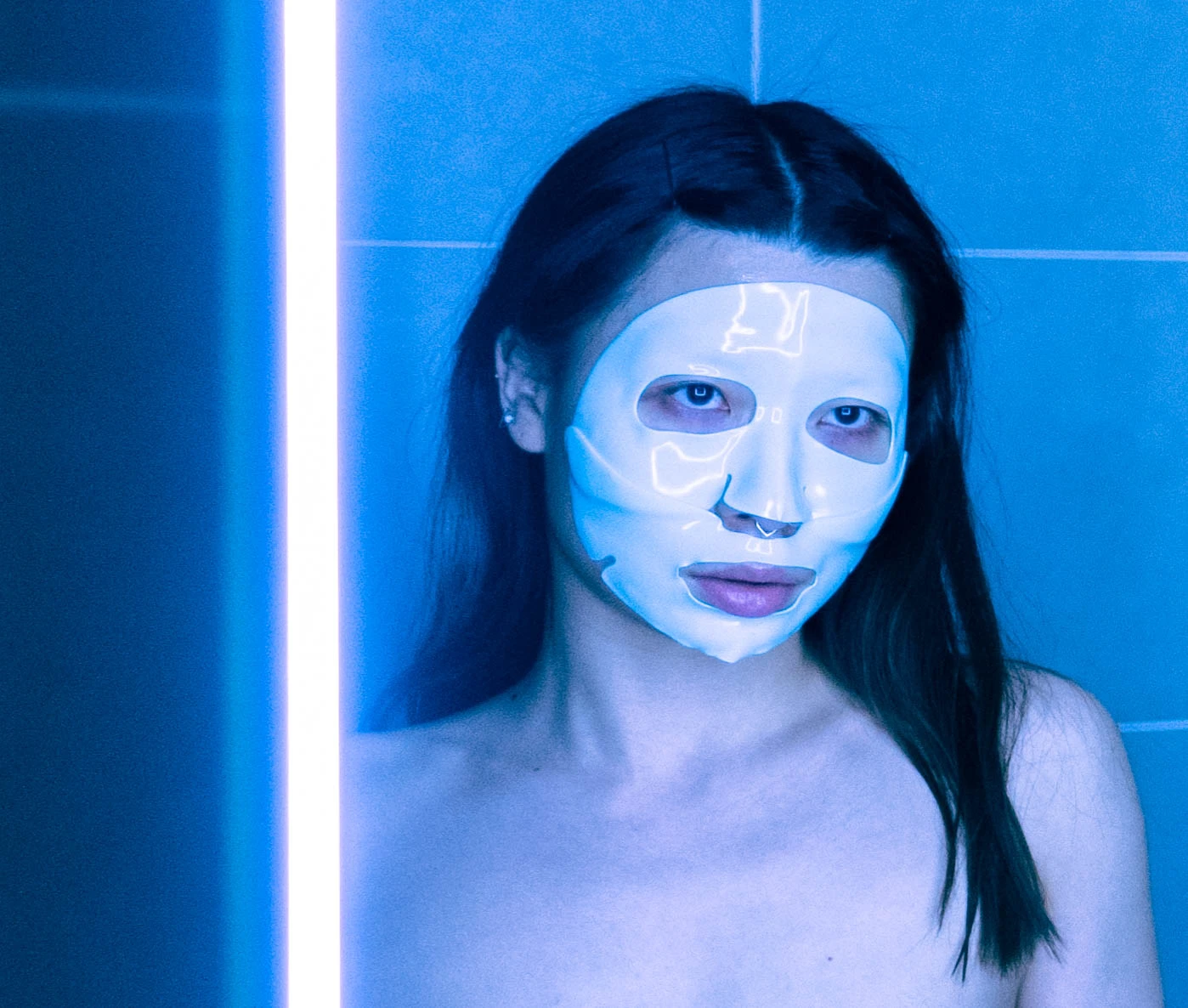 a woman wearing a rubber face mask stands in front of a neon lit mirror.