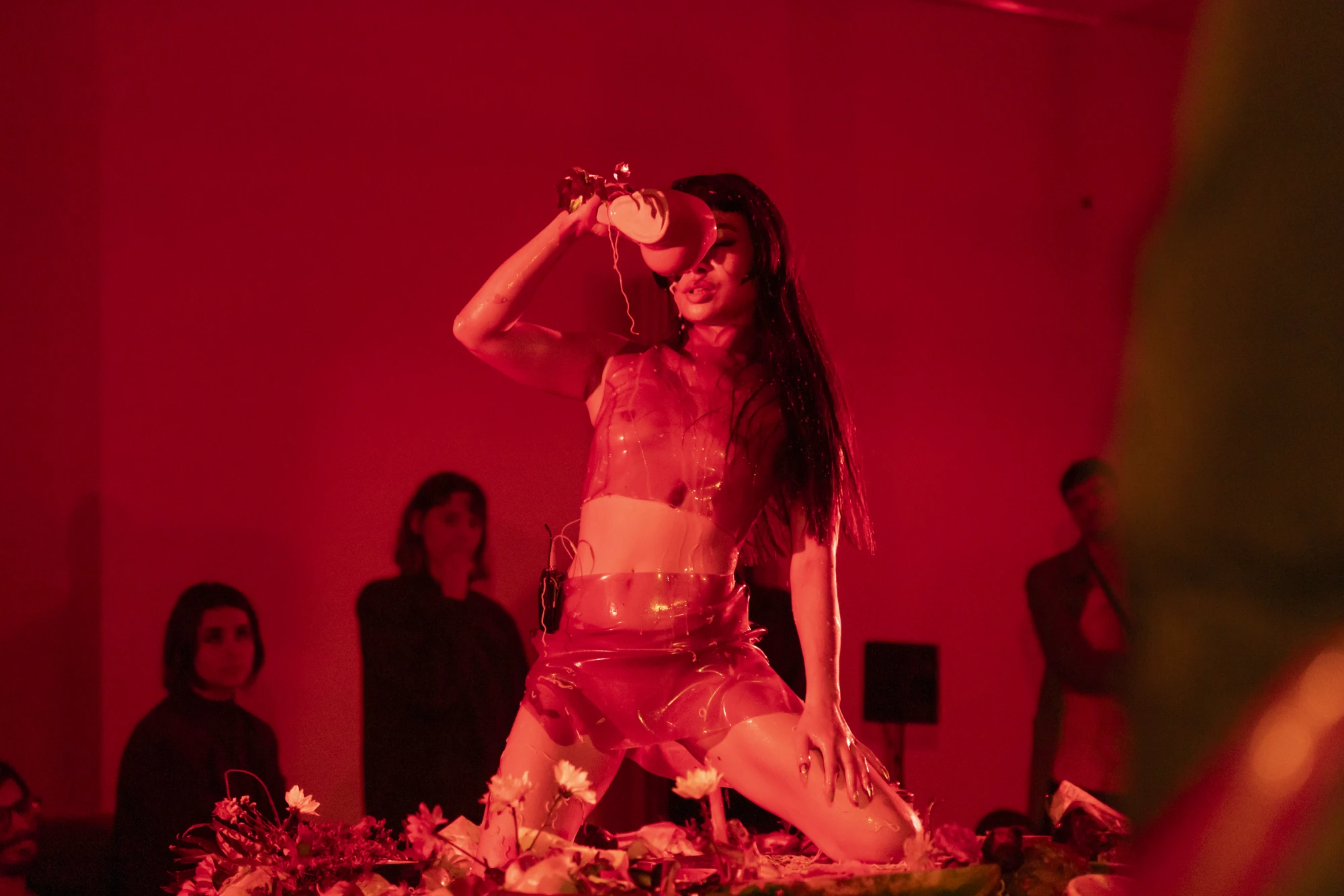 Performance documentation of a woman pourind liquid on herself. She is wearing see through latex and there is a wash of red light.