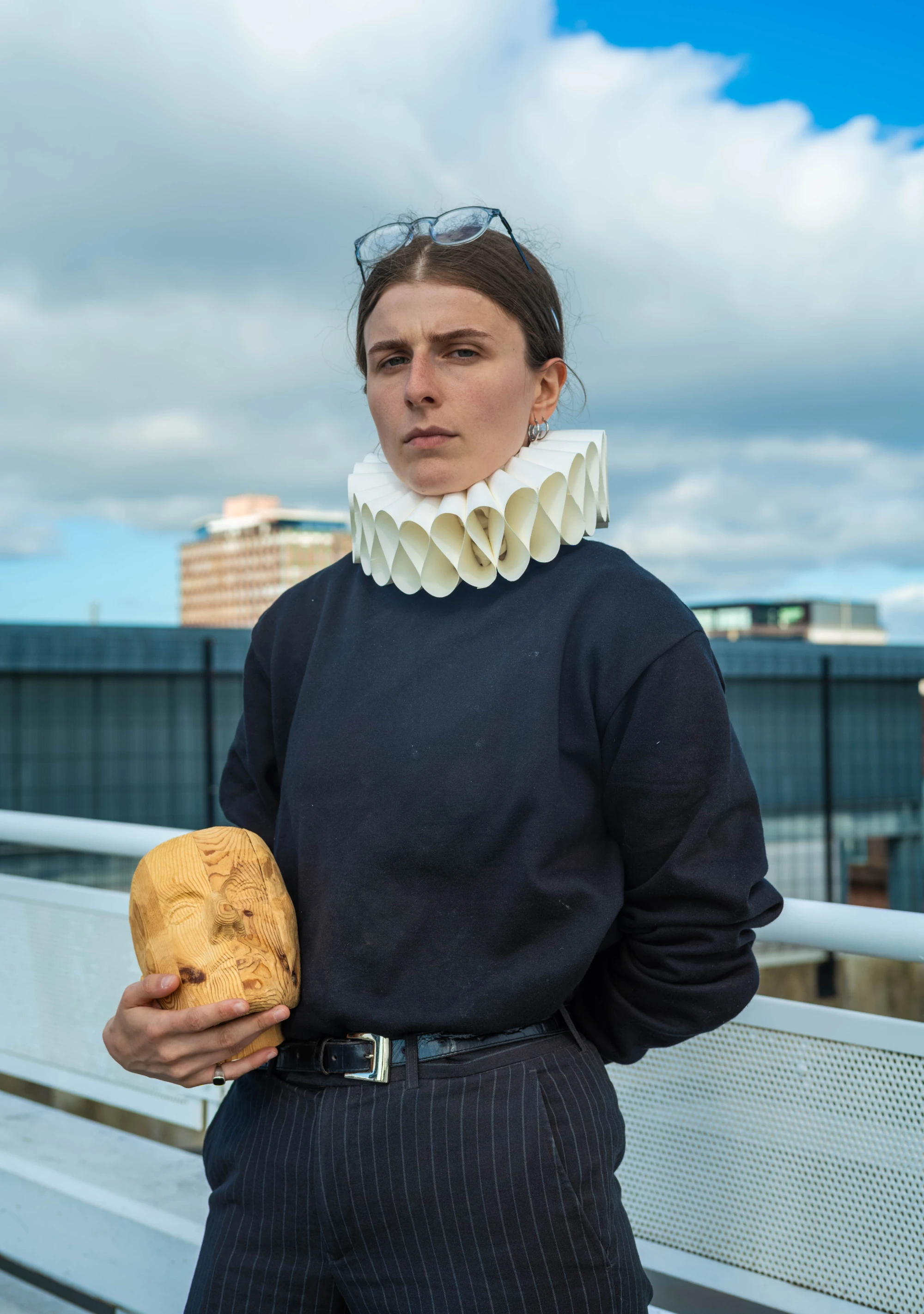 a non binary person wearing a ruff collar holds a carved wooden head.