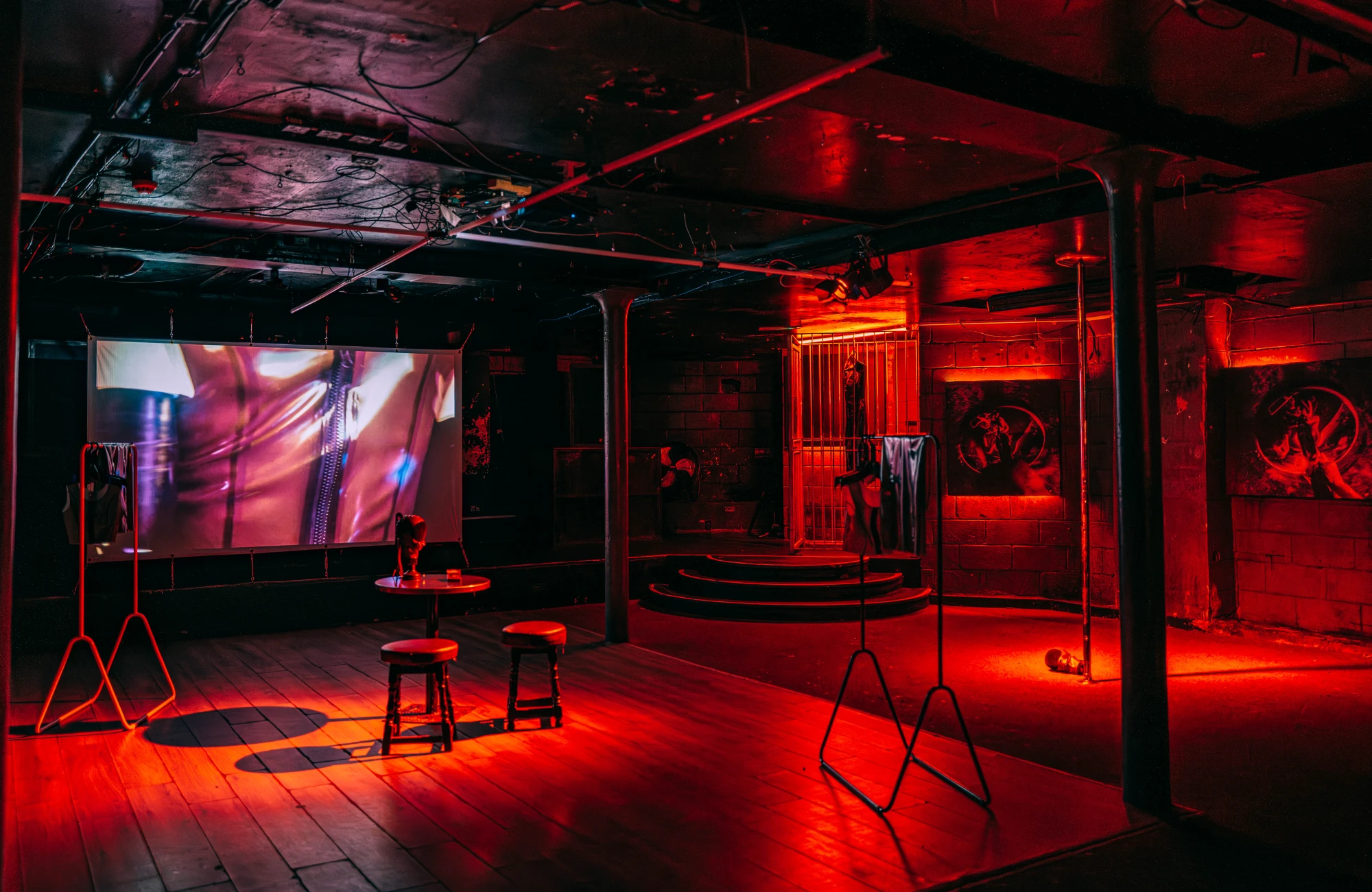 a dark red space showing a projection screen at the back, and a table with two stools at the front, with a latex dress in a cage and some photographic prints on the walls.