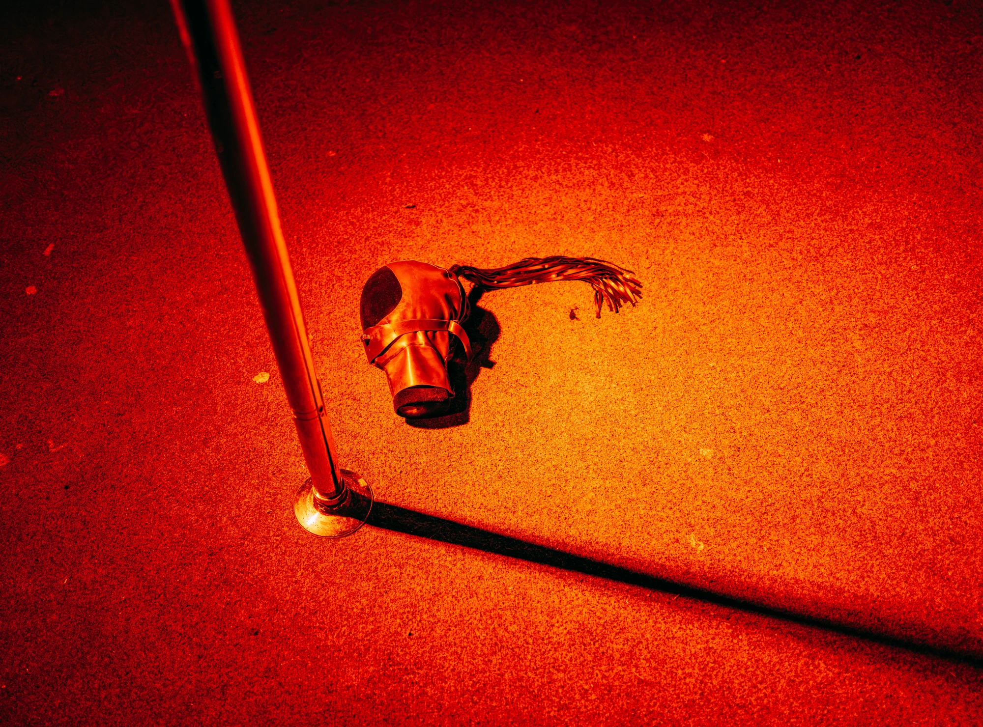 A latex mask lies on the floor next to a chrome stripper pole, under a red light.
