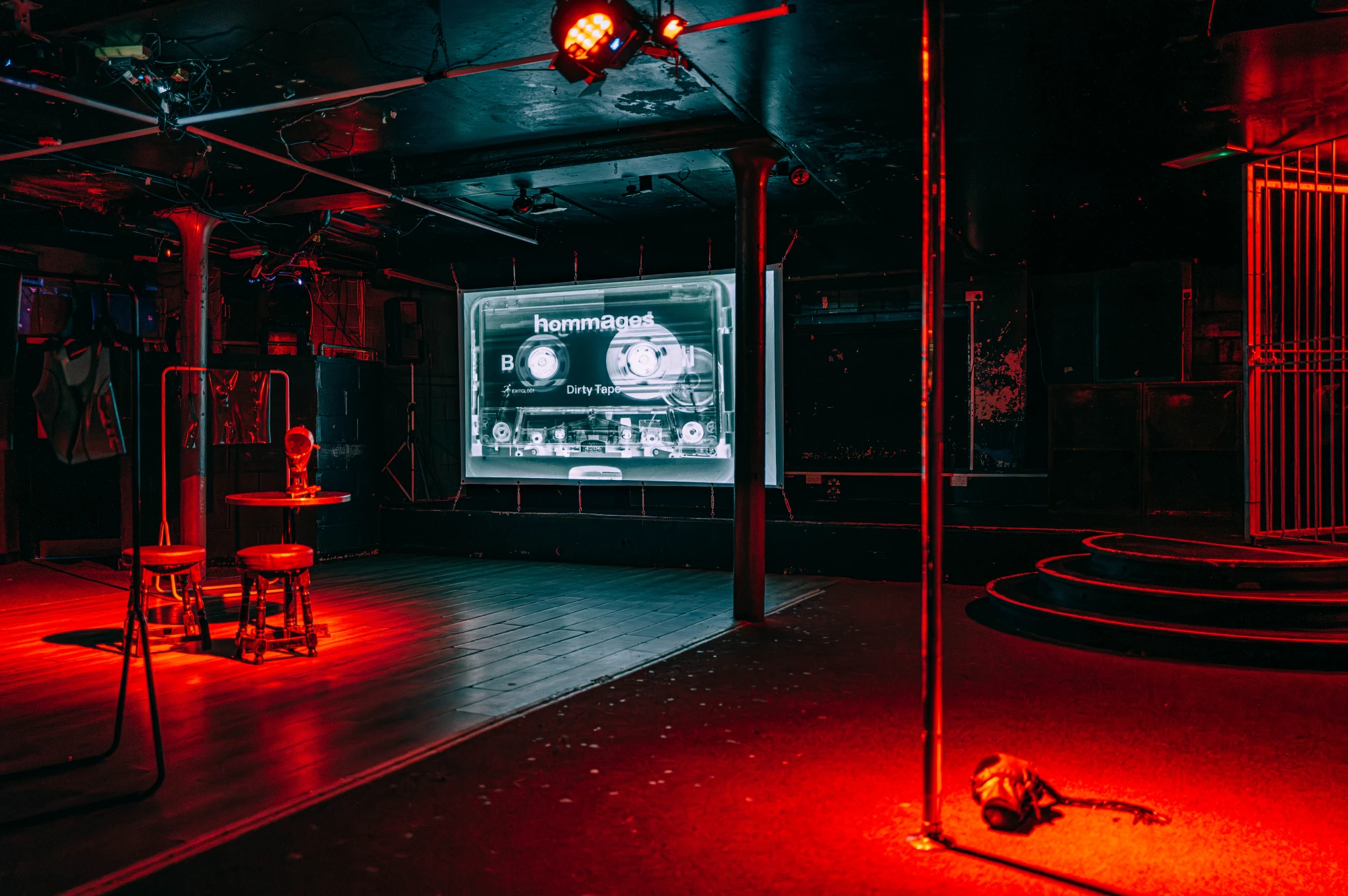 a dark red space showing a projection screen at the back, and a table with two stools at the front, and a latex hood on the floor next to a stripper pole.
