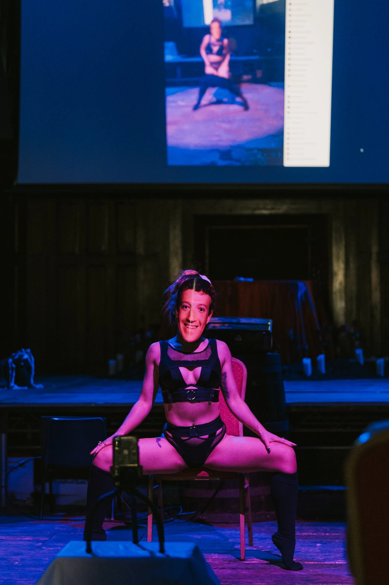 a woman wearing black hoodie and sweatpants performs as Mark Zuckerberg. The image is repeated via a big screeen above her.