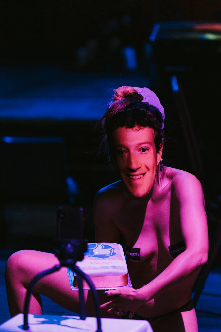 a woman wearing black hoodie and sweatpants performs as Mark Zuckerberg. She presents a cake with a like thumbs up printed on it.