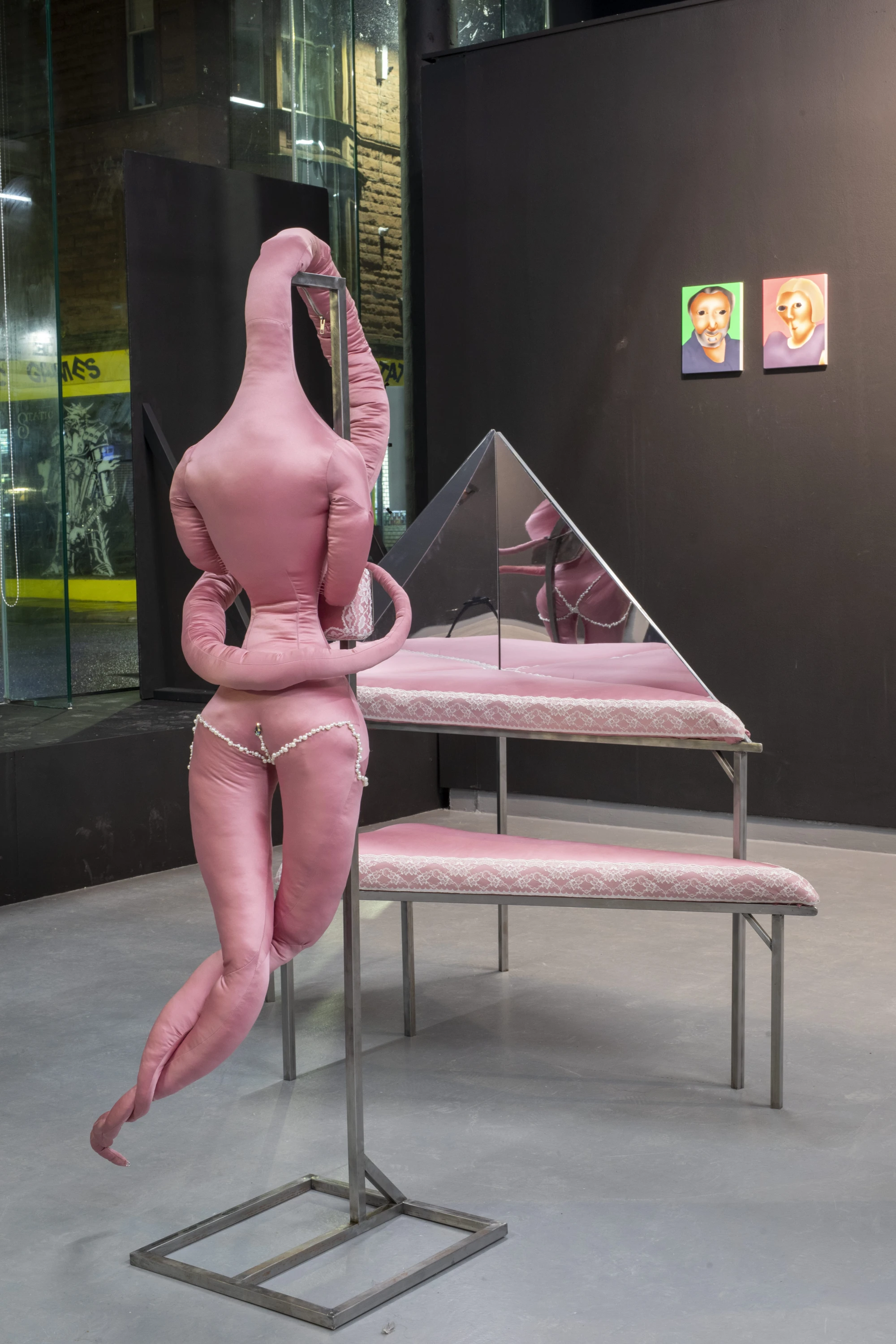 a soft pink fabric sculpture hangs on a metal stand in front of a triangular dressing table. Two paintings hang on a black wall in the back.