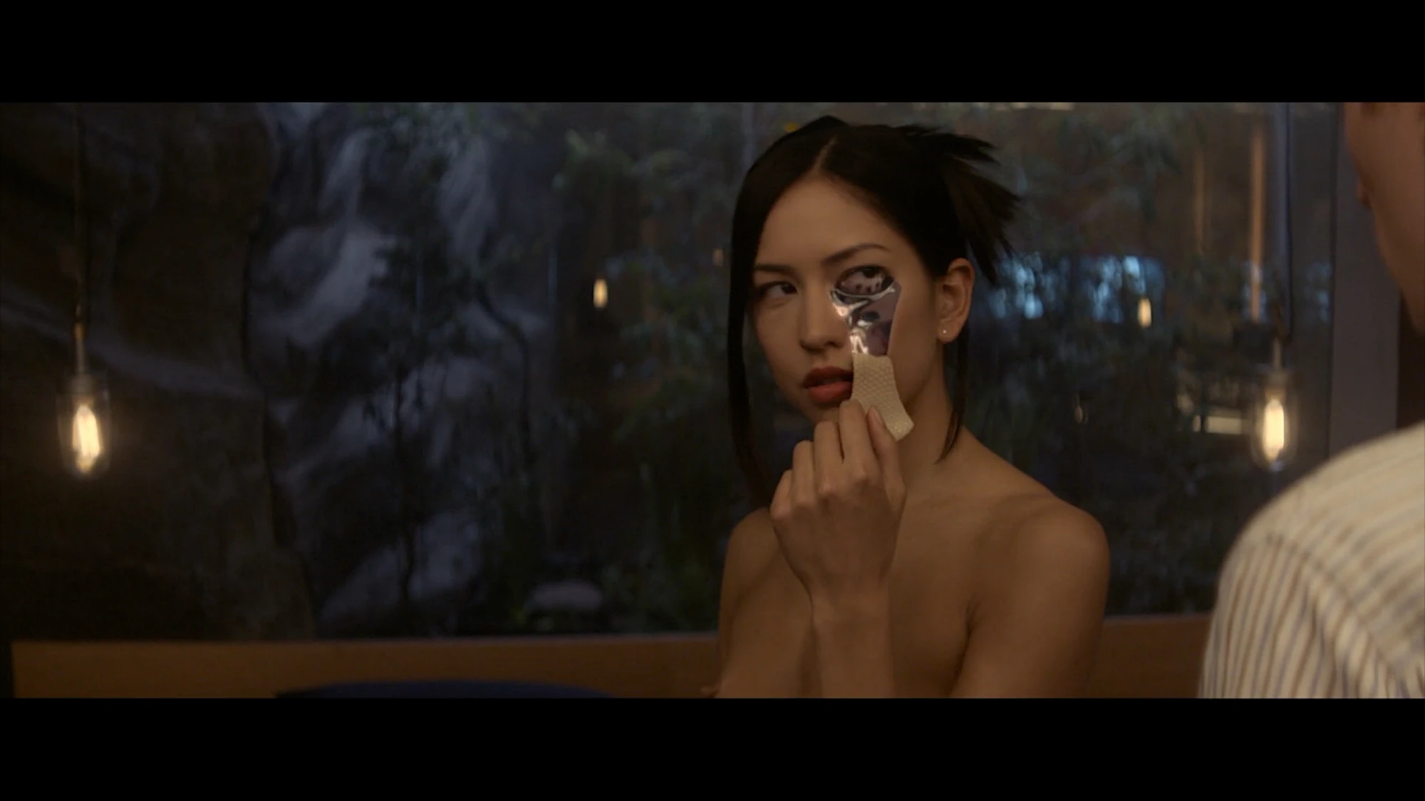 An Asian woman with bare shoulders pulls the skin off from under her eye, revealing chrome metal underneath. She's looking at a man who is not visible in the frame.