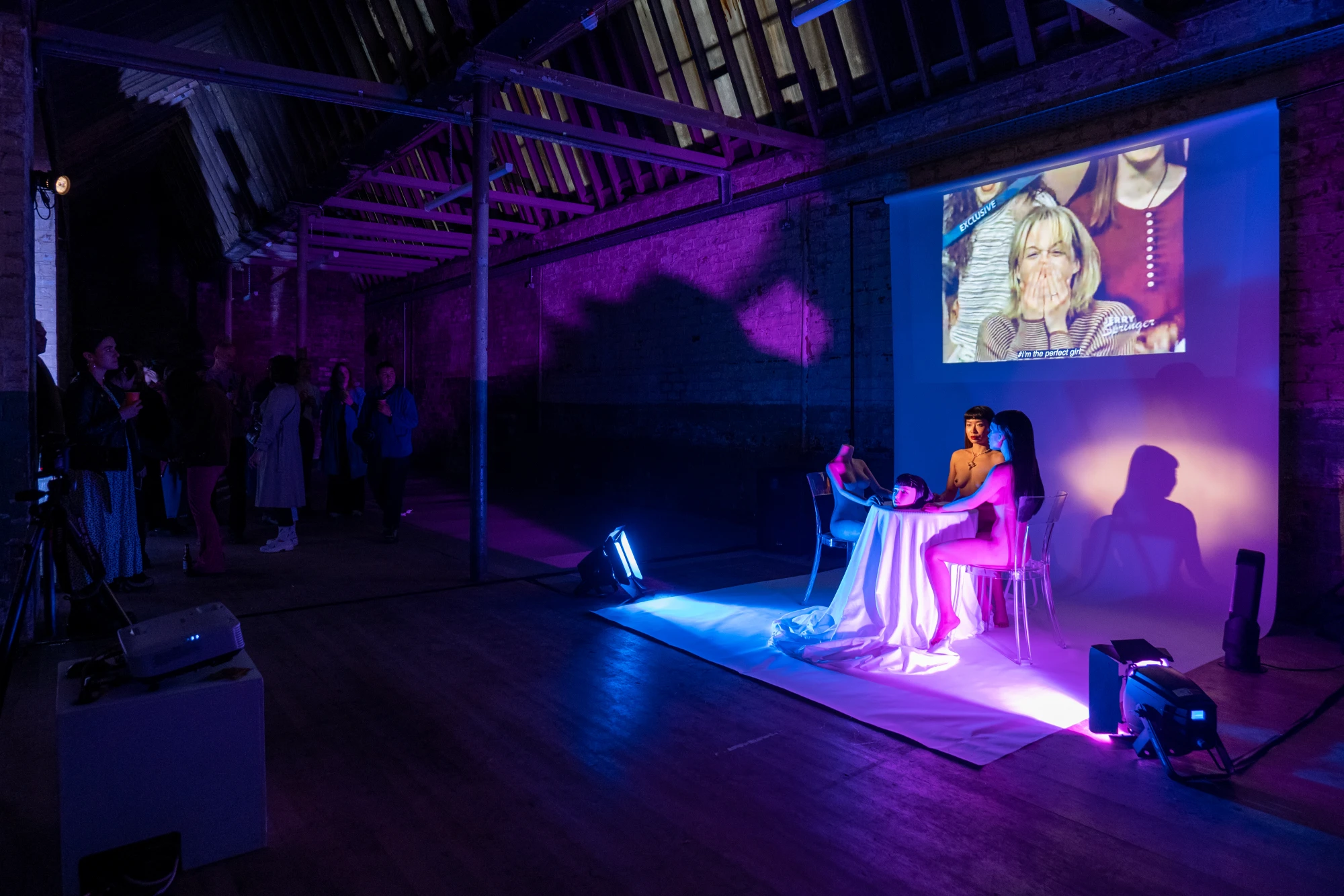 A chinese woman sits, fully naked, at a table alongside two silicone dolls, one of whom is headless. A dolls head rests on the table. Above her, a film plays on loop.