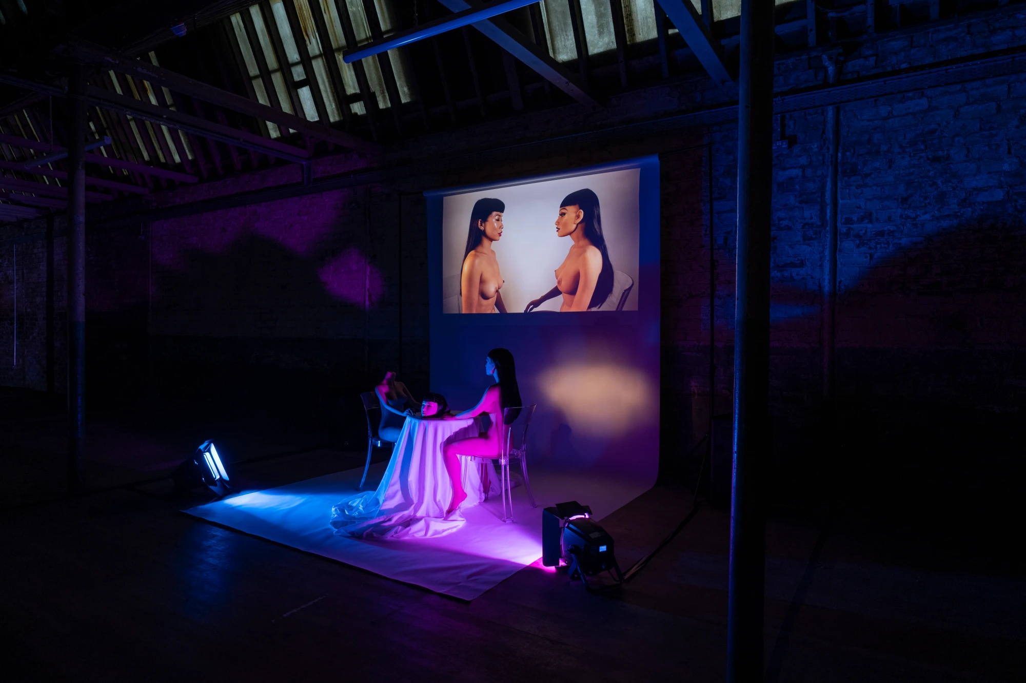 two silicone dolls sit at a table covered in white tablecloth. One of them is headless. There is a pink and blue wash of light. Above them, a film plays on loop.