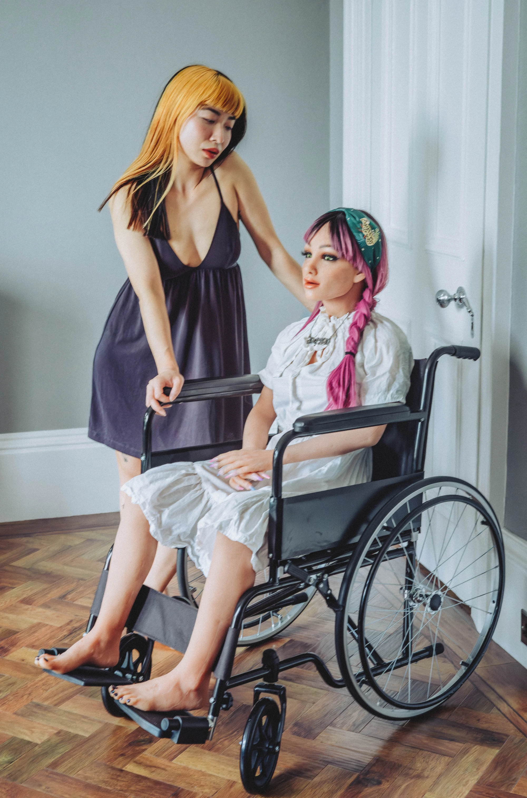 A woman stands next to a silicone love doll, who sits in a wheelchair. She is wearing a black dress and has orange hair, and the doll is wearing white with pink hair.