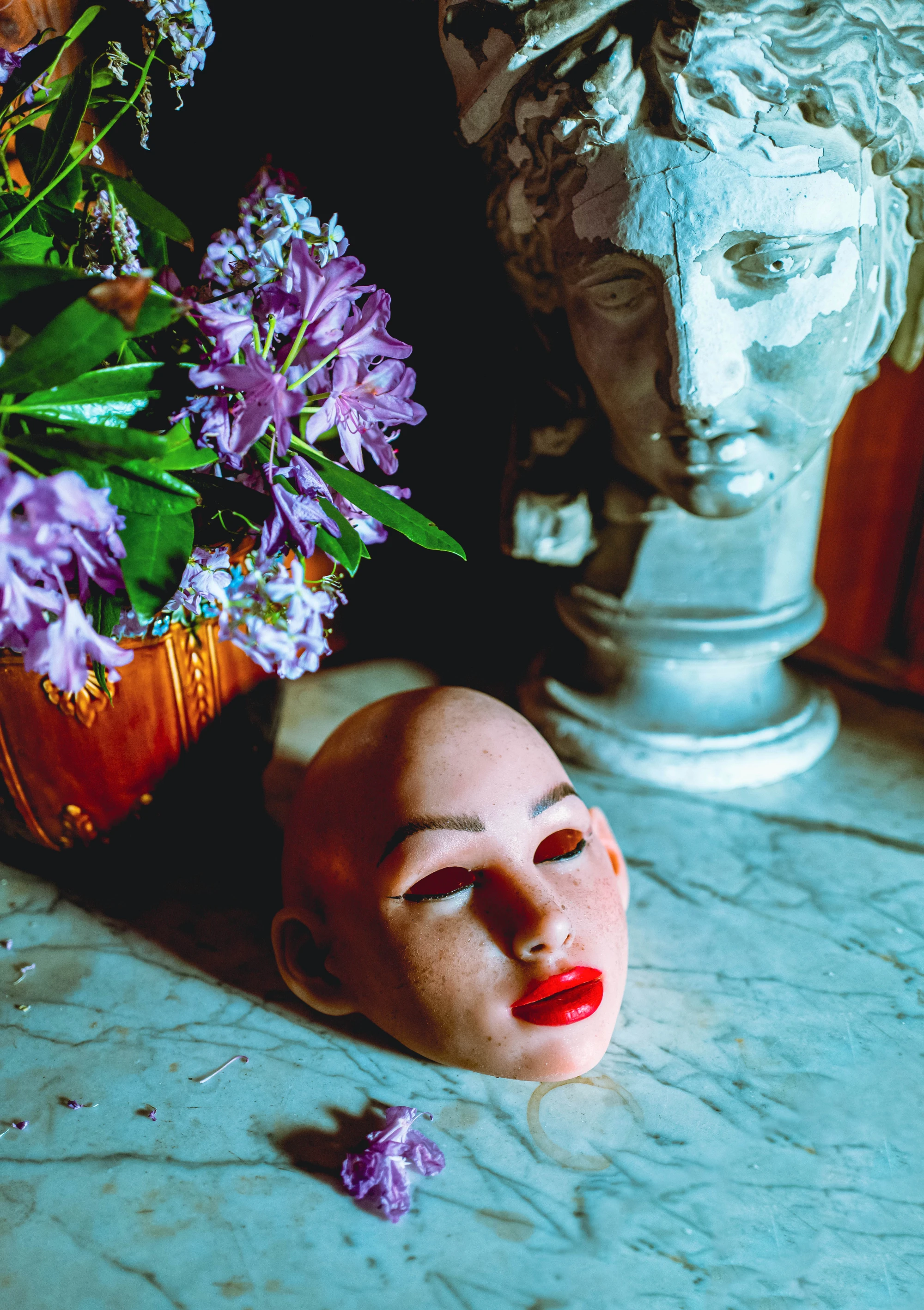 The unmounted face of a realdoll lying on top of a marble stable. A plaster cast of a head is next to it, as if observing.