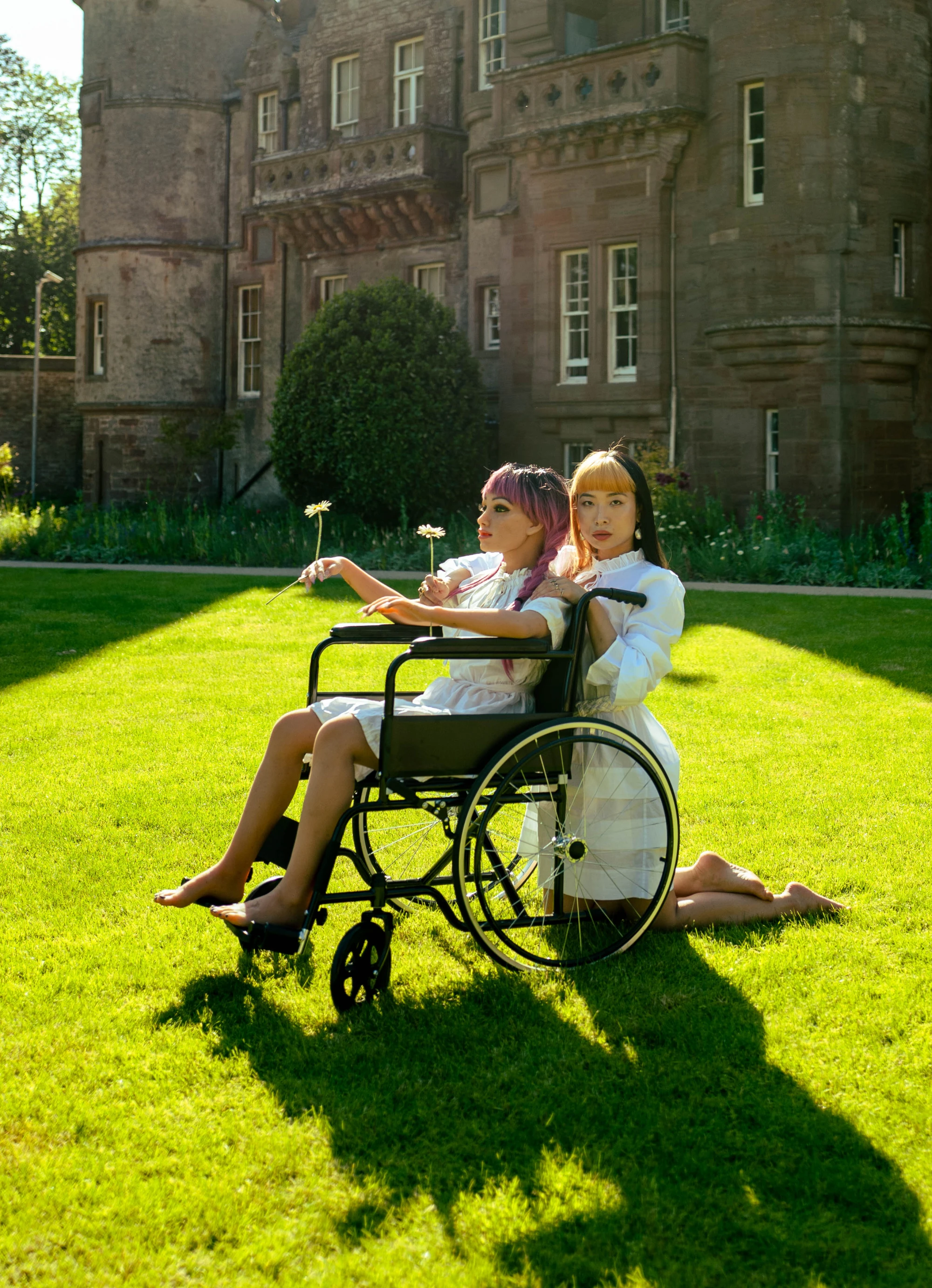 a life sized silicone love doll is in a wheelchair in front of a grand building. A woman kneels behind her. They are wearing matching white dresses.