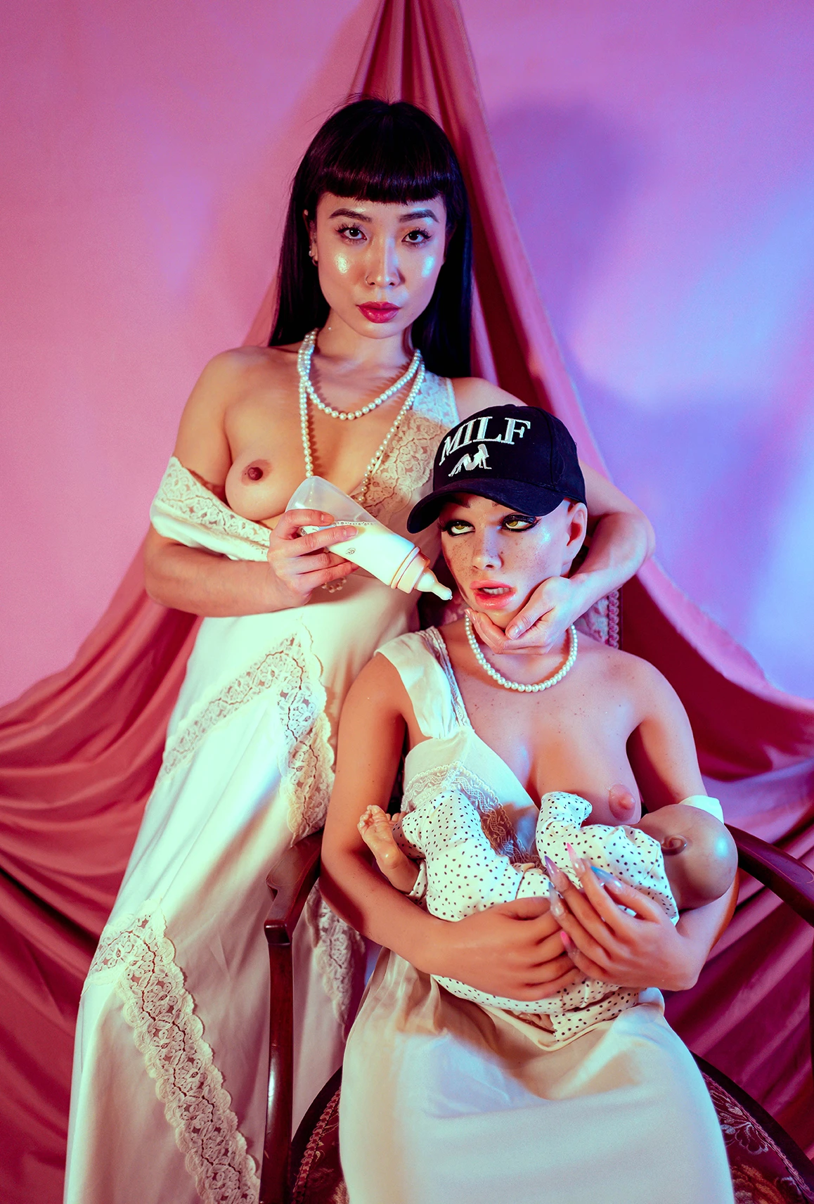 A woman stands next to a silicone love doll, she is holding a milk bottle as she feeds the doll. The doll is feeding a reborn doll whilst wearing a cap that says MILF.