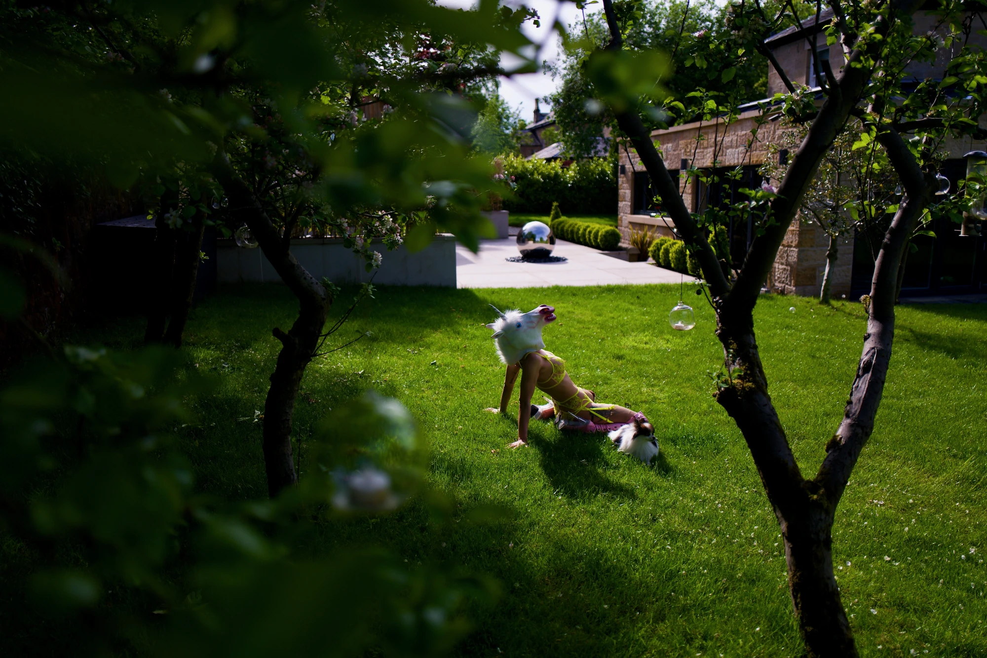 a woman with a unicorn mask and lingerie is relaxing with a pomeranian in a fancy garden.